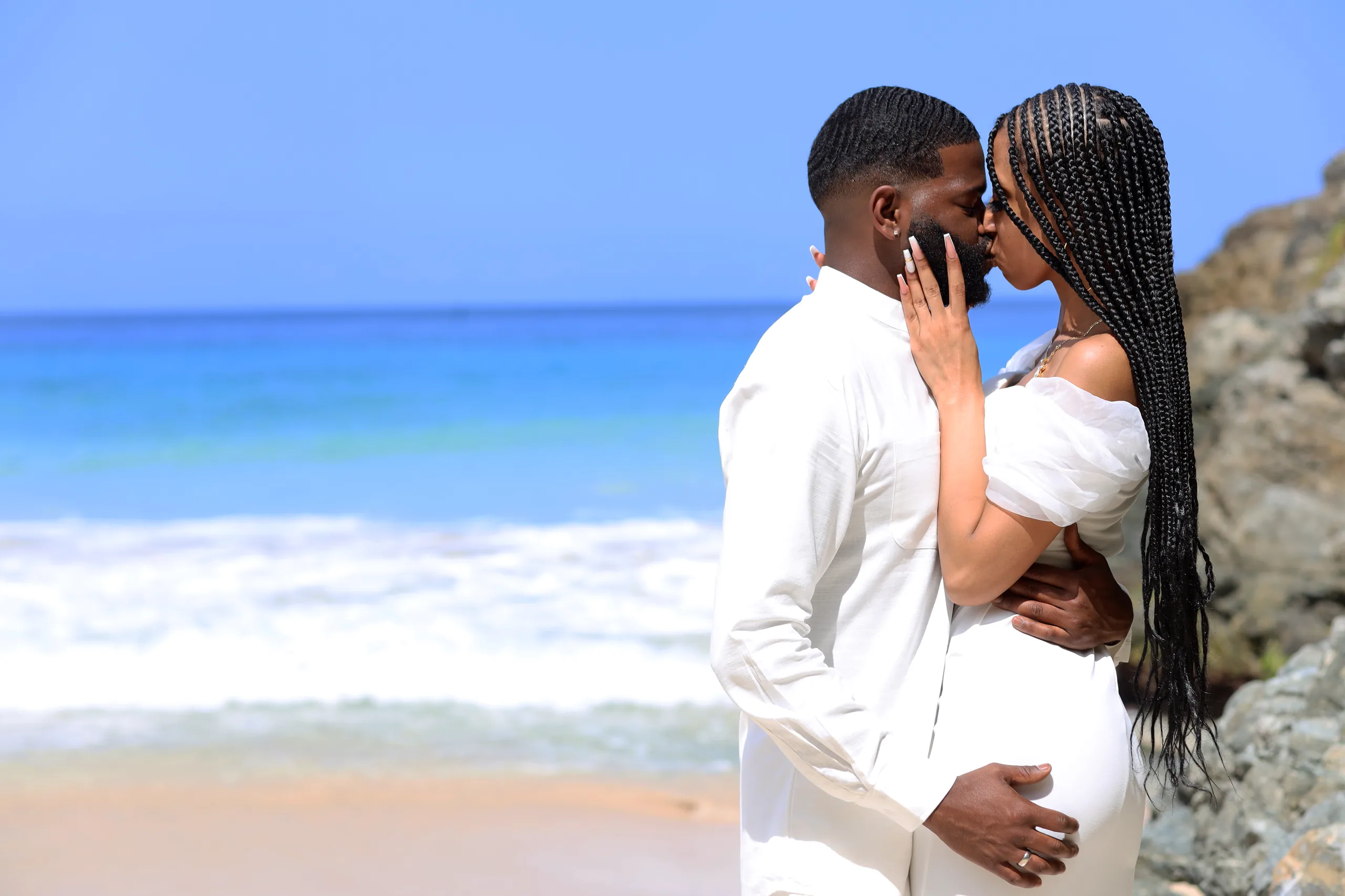 Engaged couple embracing and kissing on a rocky Caribbean beach, ocean waves in the background