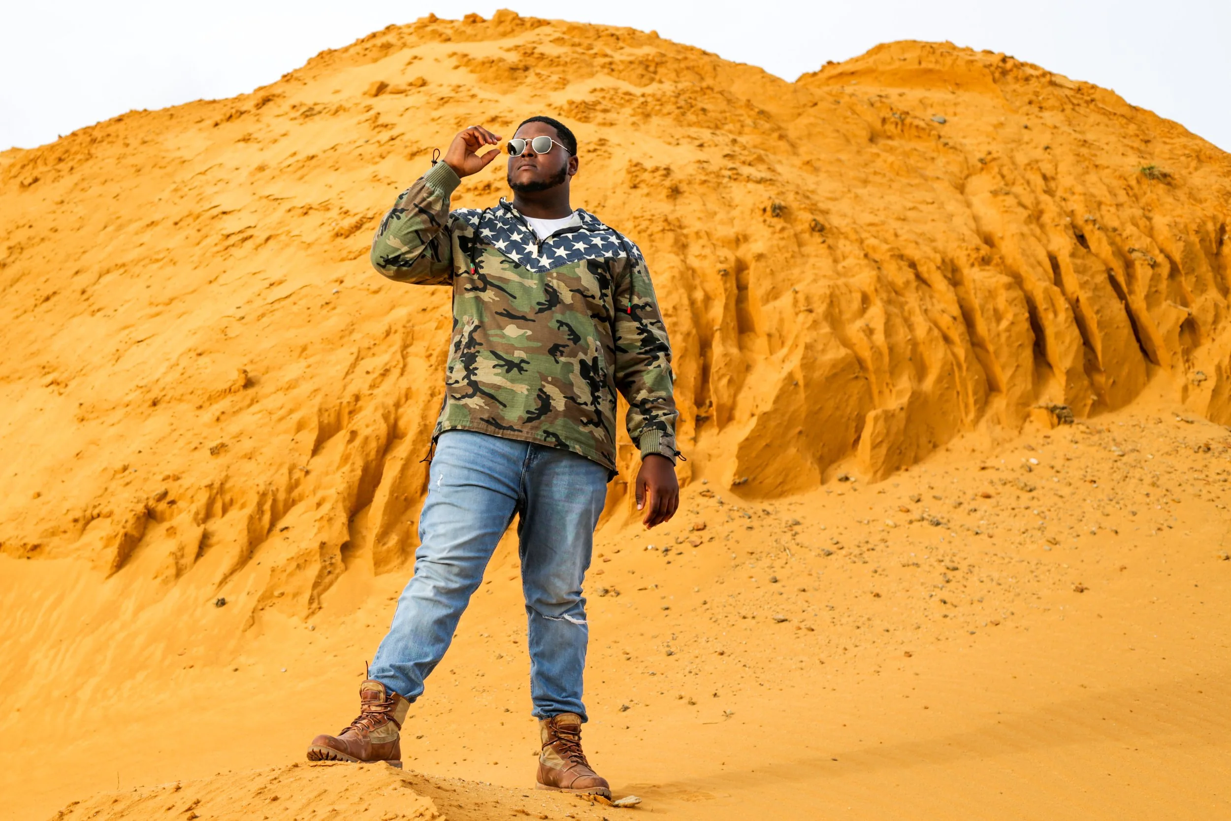 Portrait of a man in sunglasses and a camouflage jacket standing atop golden sand dunes